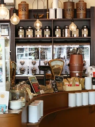 SCHAMONG KAFFEE Interior view of a stylish café featuring shelves filled with coffee and tea tins. In the foreground, there's a counter with a coffee machine and various coffee-related items.