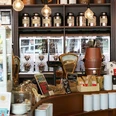SCHAMONG KAFFEE Innenansicht eines stilvollen Cafés mit Regalen voller Kaffee- und Teedosen. Im Vordergrund sind ein Tresen mit Kaffeemaschine und viele Kaffeeartikel zu sehen.Interior view of a stylish café featuring shelves filled with coffee and tea tins. In the foreground, there's a counter with a coffee machine and various coffee-related items.