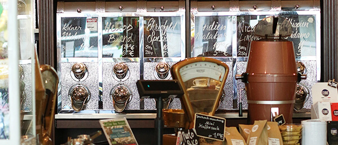 SCHAMONG KAFFEE Interior view of a stylish café featuring shelves filled with coffee and tea tins. In the foreground, there's a counter with a coffee machine and various coffee-related items.