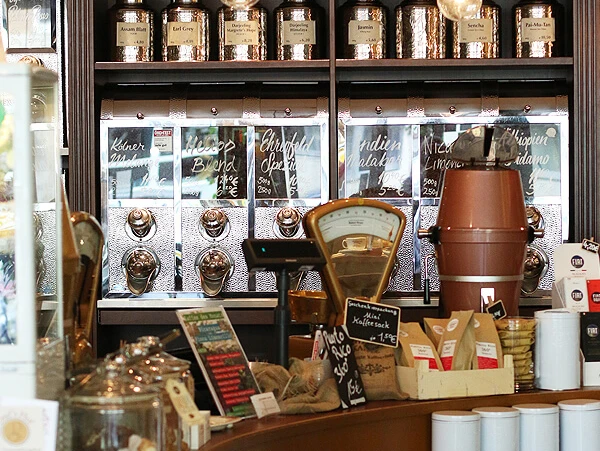 SCHAMONG KAFFEE Innenansicht eines stilvollen Cafés mit Regalen voller Kaffee- und Teedosen. Im Vordergrund sind ein Tresen mit Kaffeemaschine und viele Kaffeeartikel zu sehen.Interior view of a stylish café featuring shelves filled with coffee and tea tins. In the foreground, there's a counter with a coffee machine and various coffee-related items.