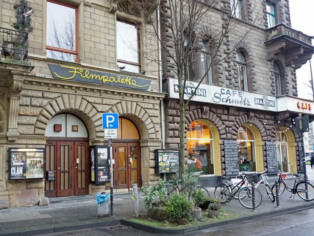 Filmpalette Ansicht des Filmtheaters "Filmpalette" in Köln, eingefasst von historischen Steingebäuden, bei grauem Wetter.The image shows the entrance of "Filmpalette", a cosy cinema located in a historic building with an ornate facade, next to a vibrant café. Bicycles are parked along the pavement, fostering a lively street atmosphere.