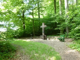 Großes Holzkreuz auf Lichtung im Wald, umgeben von Grün; nahegelegene Holzbank und Infostand.