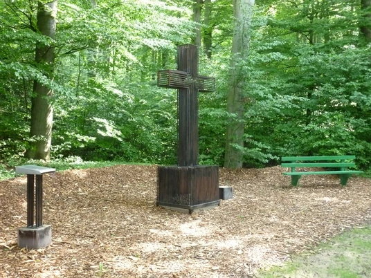 Holzkreuz in waldreicher Umgebung von Brakel-Hainhausen, flankiert von einer grünen Bank.