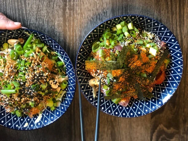Poke Makai Zwei blaue, dekorative Schalen mit üppigen Poke-Gerichten, verziert mit Frühlingszwiebeln, Nori und Sesam.