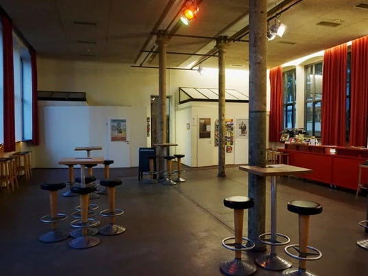 Filmhaus Im Bild des Filmhauskinos in Köln ist ein einladender, großzügiger Raum mit hoher Decke und roten Vorhängen zu sehen. Runde Hochtische mit Hockern bieten Sitzgelegenheiten, während das gedimmte Licht eine gemütliche Atmosphäre erzeugt.The picture of the Filmhaus cinema in Cologne shows an inviting, spacious room with a high ceiling and red curtains. Round high tables with stools provide seating, while the dimmed lighting creates a cozy atmosphere.