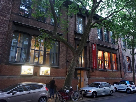 Filmhaus Das Bild zeigt eine Backstein-Fassade des Filmhauses in Köln mit großen Fenstern und Bäumen im Vordergrund.The picture shows a brick façade of the Filmhaus in Cologne with large windows and trees in the foreground.