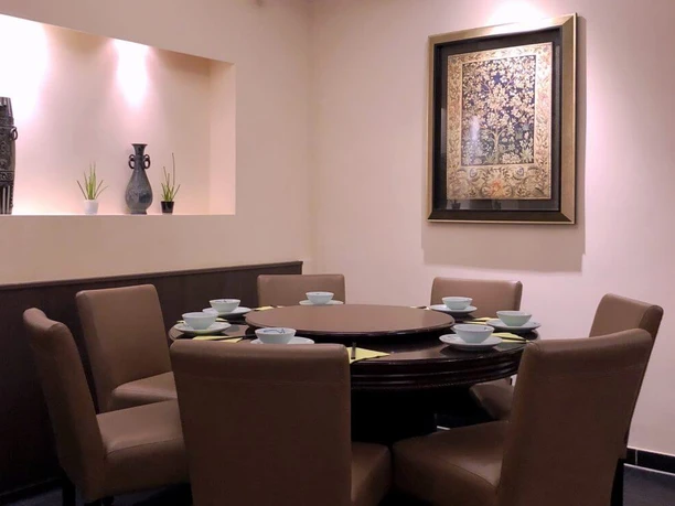 Tang Wang Ein großer runder Tisch, umgeben von gemütlichen, hellbraunen Sesseln, wird von sanftem Licht beleuchtet. Schlichte Vasen und Pflanzen in einer Nische fügen ein dezentes, dekoratives Element hinzu.