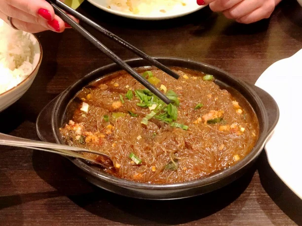Tang Wang Ein schwarzer Teller beherbergt ein köstliches Gericht aus durchscheinenden Glasnudeln, garniert mit frischen Frühlingszwiebeln. Eine Hand hält Essstäbchen. Im Hintergrund sind Reis und andere Teller zu sehen.