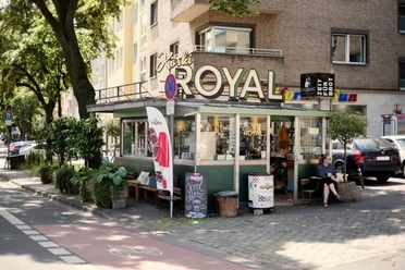 Köski Royal Ein charmanter Kiosk mit dem Schriftzug "KÖSKI ROYAL", umgeben von städtischem Grün in einer urbanen Umgebung.