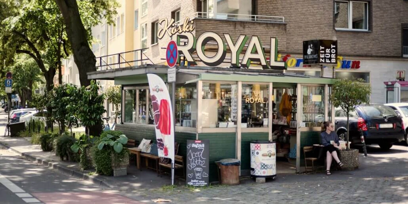 Köski Royal Ein charmanter Kiosk mit dem Schriftzug "KÖSKI ROYAL", umgeben von städtischem Grün in einer urbanen Umgebung.A charming kiosk with the lettering "KÖSKI ROYAL", surrounded by urban greenery in an urban environment.
