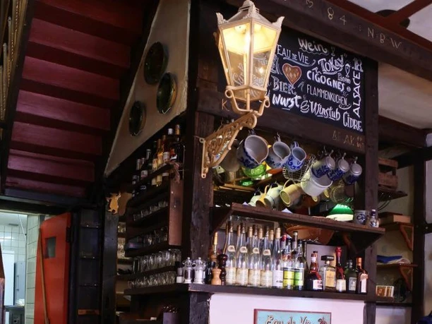 Wackes Ein gemütlicher Gastraum mit rot-weiß karierten Tischdecken, Holzregalen voller Wein und Getränken sowie einer stilvollen Lampe, die den Raum erhellt.