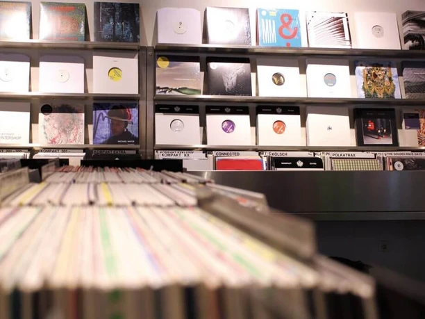 Kompakt The picture shows a stylishly furnished record store. In the foreground, numerous records can be seen arranged on shelves, representing a variety of musical genres.