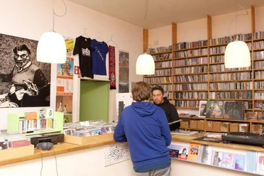 A-Musik Im Vordergrund steht eine Person, die mit einem Verkäufer spricht, während im Hintergrund Regale voller Vinyl-Schallplatten den Raum zieren. Licht strahlt von modernen Deckenlampen, und an der Wand hängt ein großes Schwarz-Weiß-Poster.