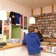 A-Musik Im Vordergrund steht eine Person, die mit einem Verkäufer spricht, während im Hintergrund Regale voller Vinyl-Schallplatten den Raum zieren. Licht strahlt von modernen Deckenlampen, und an der Wand hängt ein großes Schwarz-Weiß-Poster.In the foreground is a person talking to a salesperson, while shelves full of vinyl records adorn the room in the background. Light shines from modern ceiling lamps and a large black and white poster hangs on the wall.
