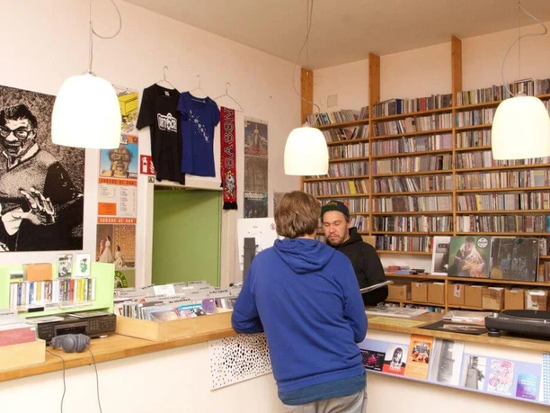 A-Musik In the foreground is a person talking to a salesperson, while shelves full of vinyl records adorn the room in the background. Light shines from modern ceiling lamps and a large black and white poster hangs on the wall.