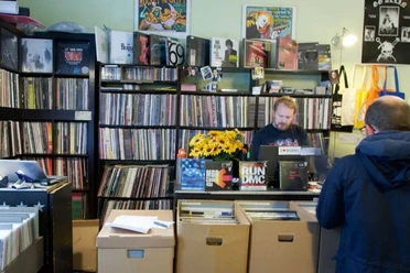 Underdog Recordstore In einem kleinen Plattenladen stehen eng gestellte Regale voller Schallplatten. Ein Verkäufer, umgeben von bunten Plattencovern und Postern an den Wänden, steht hinter einer Theke mit Kasse. Vor der Theke befinden sich Kartons mit weiteren Schallplatten.