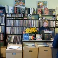 Underdog Recordstore In einem kleinen Plattenladen stehen eng gestellte Regale voller Schallplatten. Ein Verkäufer, umgeben von bunten Plattencovern und Postern an den Wänden, steht hinter einer Theke mit Kasse. Vor der Theke befinden sich Kartons mit weiteren Schallplatten.In a small record store, shelves are crammed full of records. A sales assistant, surrounded by colorful record covers and posters on the walls, stands behind a counter with a cash register. In front of the counter are boxes with more records.