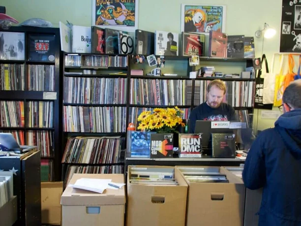 Underdog Recordstore In a small record store, shelves are crammed full of records. A sales assistant, surrounded by colorful record covers and posters on the walls, stands behind a counter with a cash register. In front of the counter are boxes with more records.