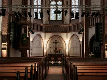 Kulturkirche Innenansicht der Kulturkirche Köln mit Holzbankreihen, erhöhter Bühne und buntglasverzierten Fenstern.Interior view of the Kulturkirche Köln with rows of wooden benches, raised stage and stained glass windows.