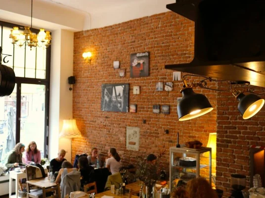 Wohnraum Gemütliches Café mit Backsteinwand, großen Fenstern und geselligen Menschen an Tischen.Cozy café featuring brick walls, large windows, and sociable guests seated around tables.
