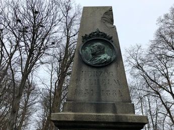Gedenksäule mit Relief von Herzog Wilhelm, umgeben von winterlichen Bäumen unter grauem Himmel.