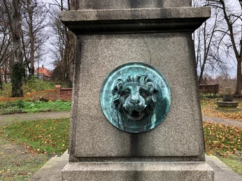 Bronzerelief eines Löwenkopfes an einem steinernen Denkmal in herbstlicher Parklandschaft.