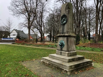 Herzog-Wilhelm Denkmal in einem grünen Park, umgeben von herbstlichen Bäumen und Gebäuden im Hintergrund.