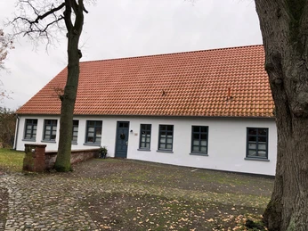Küsterhaus Daverden Ein traditionelles Gebäude mit rotem Ziegeldach und mehreren blauen Fenstern, umgeben von Bäumen.