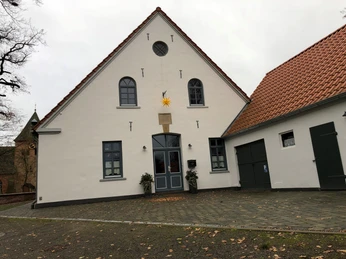 Küsterhaus Daverden Weißes, zweigeschossiges Gebäudemit Satteldach und Zierstern über der Tür, umgeben von Laub und Bäumen.