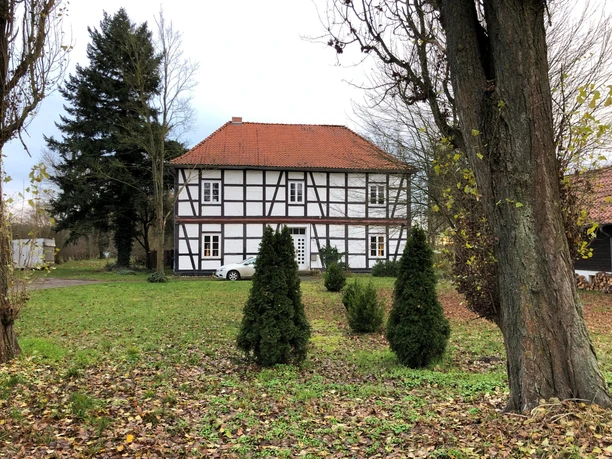 Das Bild zeigt ein historisches Fachwerkhaus im Grünen mit kleinen Koniferen im Vordergrund.