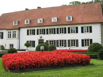 Blumenbeet vor dem Haus des Gastes Blumenbeet mit leuchtend roten Blumen vor einem historischen Gebäude mit weißer Fassade und roten Dach.