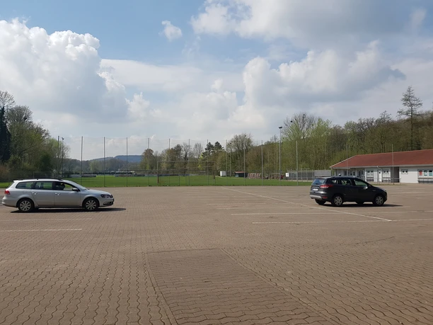 Parkplatz am Sportplatz Bad Holzhausen Leerer, gepflasterter Parkplatz vor einem begrünten Sportplatz, zwei geparkte Autos, bewölkter Himmel.