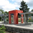 Ein moderner Bahnsteig mit Glasunterständen und einer roten Überdachung in ländlicher Umgebung.
