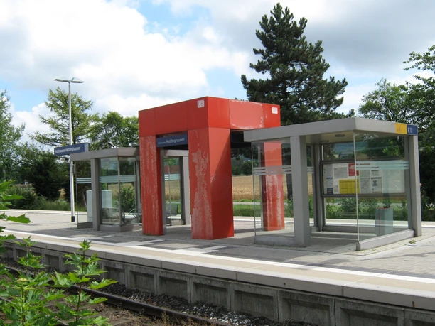 Ein moderner Bahnsteig mit Glasunterständen und einer roten Überdachung in ländlicher Umgebung.