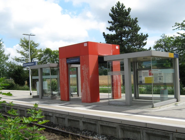 Ein moderner Bahnsteig mit Glasunterständen und einer roten Überdachung in ländlicher Umgebung.