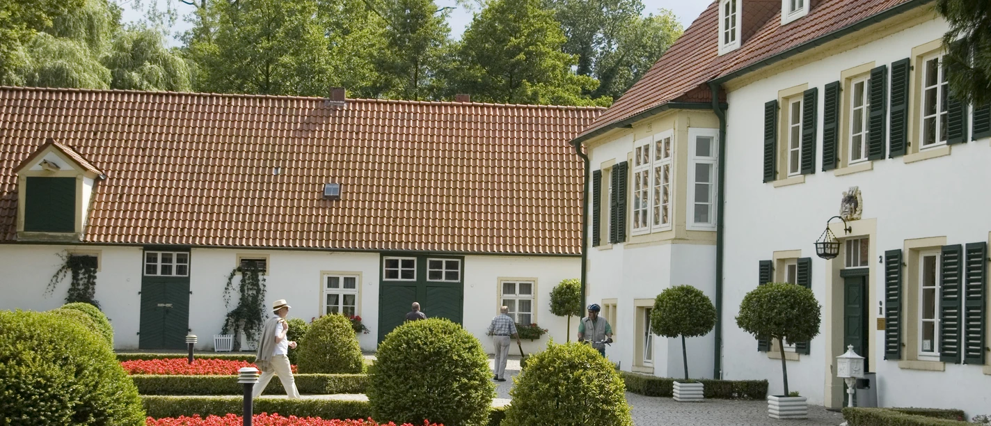 Haus des Gastes im Sommer Weiße Gebäude mit rotem Ziegeldach, umgeben von gepflegten Beeten und Bäumen im Sommer.