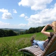 Landschaftsliege in Niese Frau genießt von einer Holzliege aus den weiten Blick über grüne Hügel und blauen Himmel.