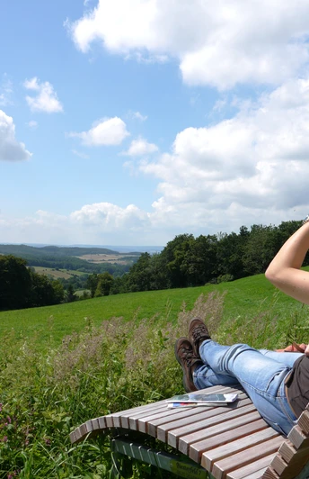 Landschaftsliege in Niese Frau genießt von einer Holzliege aus den weiten Blick über grüne Hügel und blauen Himmel.
