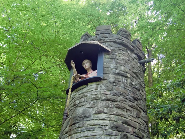 Deutscher Märchenwald Altenberg Figur der Rapunzel zieht Zopf aus Fenster eines steinernen Turms im grünen, bewaldeten Umfeld.