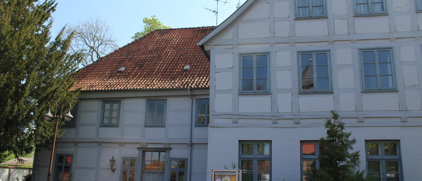 Historisches weißes Fachwerkgebäude mit rotem Ziegeldach, bunten Fenstern und grünem Baum im Vordergrund.