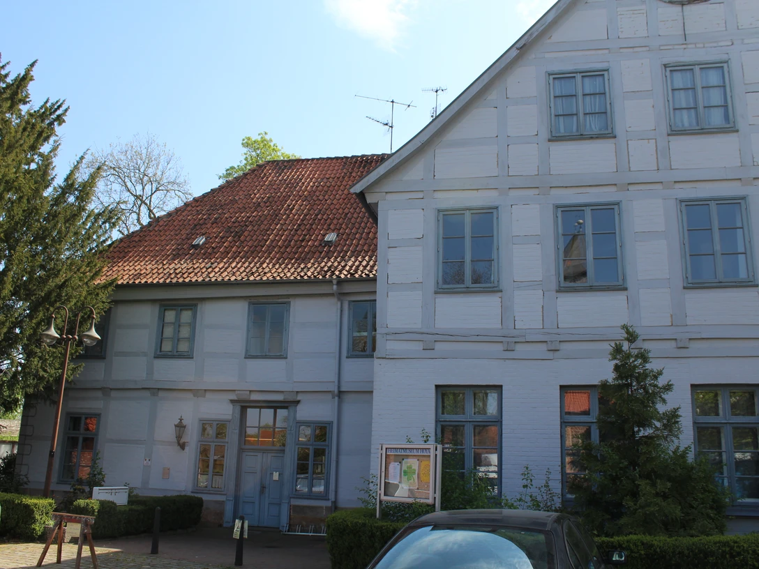 SG Grafschaft Hoya Heimatmuseum Historisches weißes Fachwerkgebäude mit rotem Ziegeldach, bunten Fenstern und grünem Baum im Vordergrund.