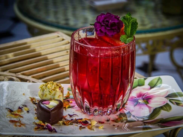 Seiberts Cocktailbar A red beverage served in a stylish glass, garnished with a purple flower, a strawberry, and mint, placed on a plate decorated with artistic pralines and flower petals.