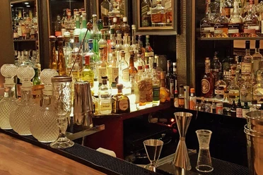 Ona Mor Cocktailbar Auf dem Bild ist eine elegante Bar mit einer Vielzahl von dekorativ beleuchteten Spirituosenflaschen, kunstvoll gestalteten Gläsern und Barutensilien auf einem edlen Holztresen zu sehen, was eine gemütliche und stilvolle Atmosphäre vermittelt.