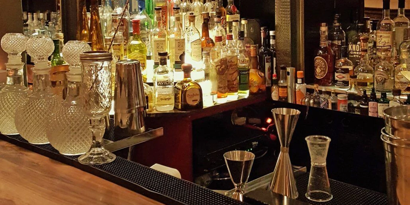 Ona Mor Cocktailbar Auf dem Bild ist eine elegante Bar mit einer Vielzahl von dekorativ beleuchteten Spirituosenflaschen, kunstvoll gestalteten Gläsern und Barutensilien auf einem edlen Holztresen zu sehen, was eine gemütliche und stilvolle Atmosphäre vermittelt.The image shows an elegant bar with a variety of decoratively lit spirits bottles, artistically designed glasses and bar utensils on a fine wooden counter, creating a cosy and stylish atmosphere.