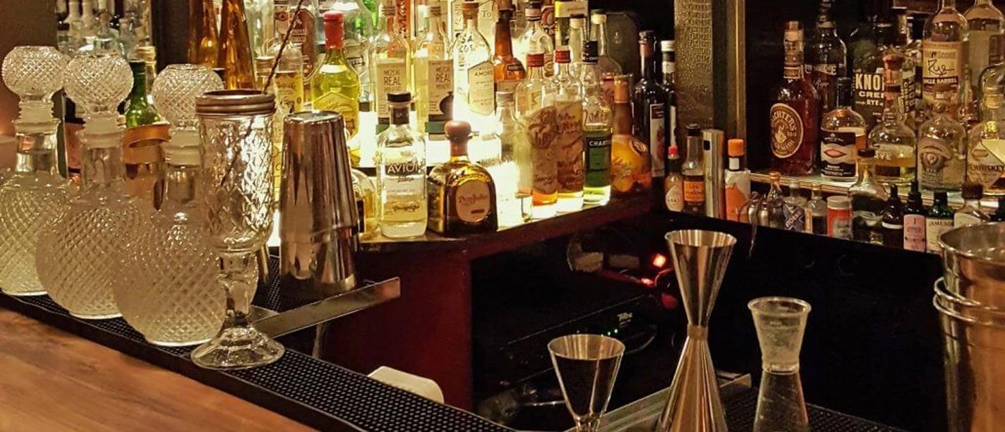 Ona Mor Cocktailbar The image shows an elegant bar with a variety of decoratively lit spirits bottles, artistically designed glasses and bar utensils on a fine wooden counter, creating a cosy and stylish atmosphere.