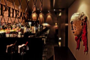 Samuel's Shep Eine stilvolle Bar mit einer kunstvollen Maske an der Wand. Die Bar ist elegant beleuchtet und lädt zu einem entspannten Abend ein.