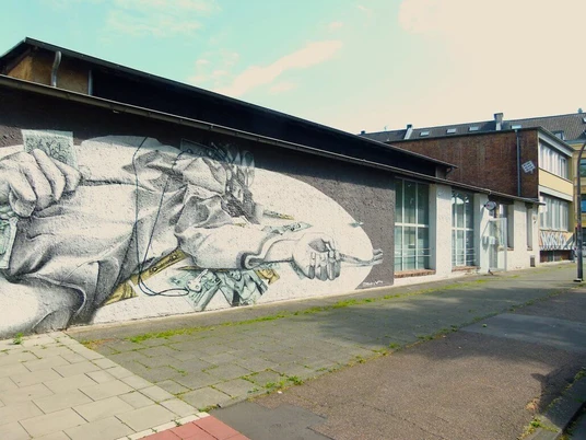 artrmx e. V. | Atelierzentrum Ehrenfeld Auf einer grauen Wand befindet sich ein beeindruckendes Wandbild. Es zeigt eine detailreiche Zeichnung eines aus Papier bestehenden Schiffes, das durch eingebettete Geldnoten segelt. Die Umgebung ist urban und ruhig, mit angrenzenden Gebäuden im Hintergrund.There is an impressive mural on a gray wall. It shows a detailed drawing of a ship made of paper sailing through embedded banknotes. The surroundings are urban and calm, with adjacent buildings in the background.