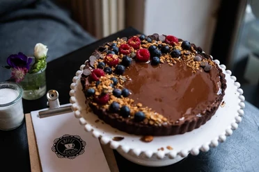 Cafe Adeli Schokoladenkuchen mit Beeren und Streuseln auf Tortenteller, im Hintergrund Blumen und Menükarte.