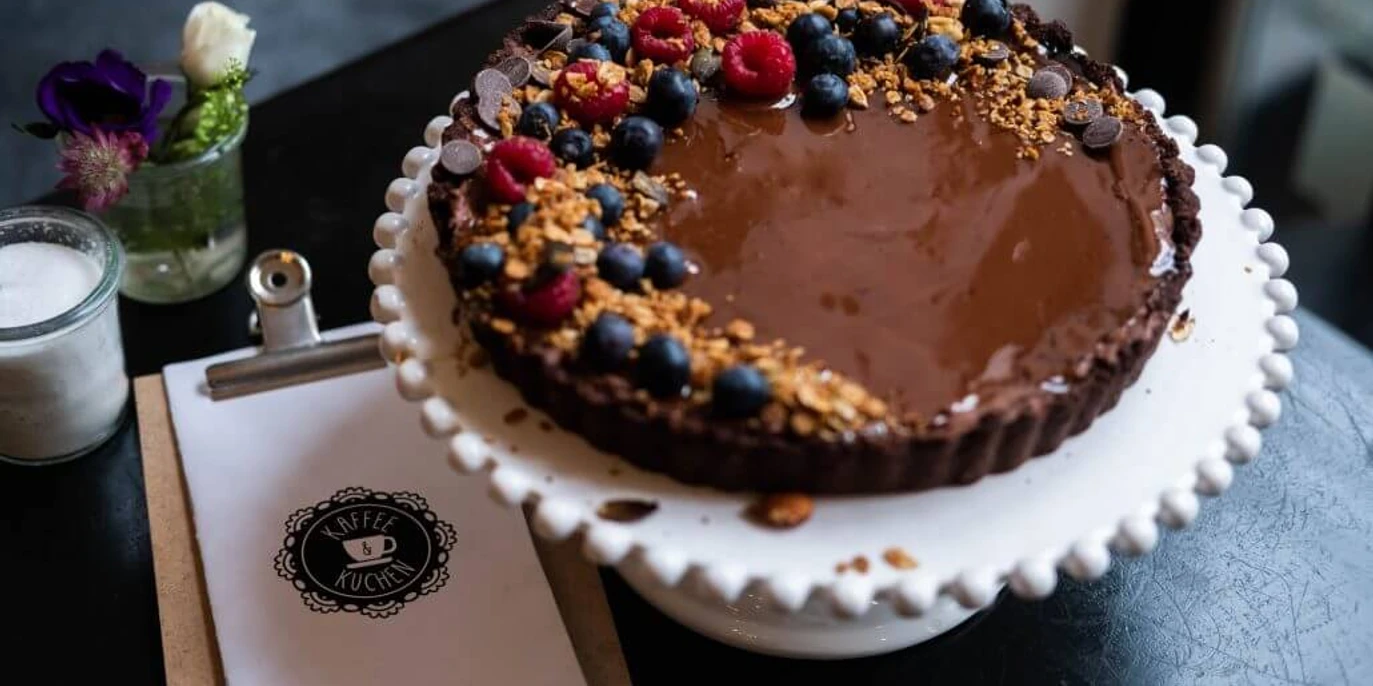 Cafe Adeli Schokoladenkuchen mit Beeren und Streuseln auf Tortenteller, im Hintergrund Blumen und Menükarte.Chocolate cake with berries and sprinkles on a cake plate, flowers and menu card in the background.