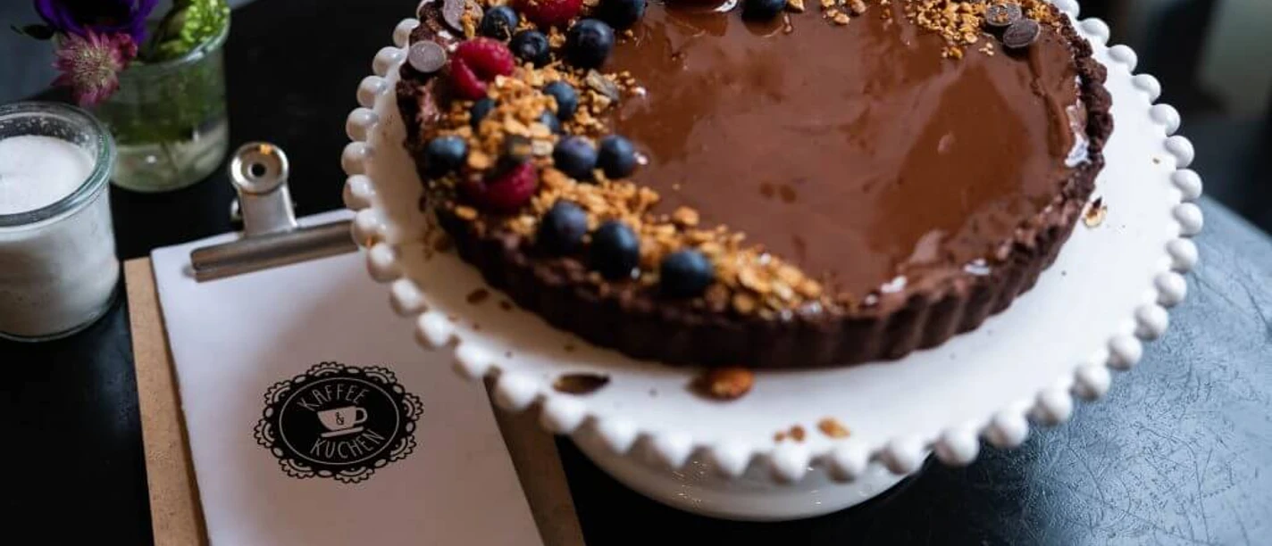 Cafe Adeli Schokoladenkuchen mit Beeren und Streuseln auf Tortenteller, im Hintergrund Blumen und Menükarte.